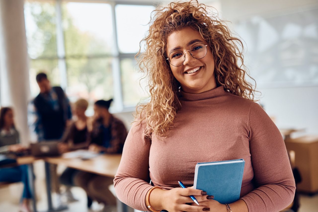 portrait-of-happy-female-student-at-college-classroom-looking-at-camera-.jpg